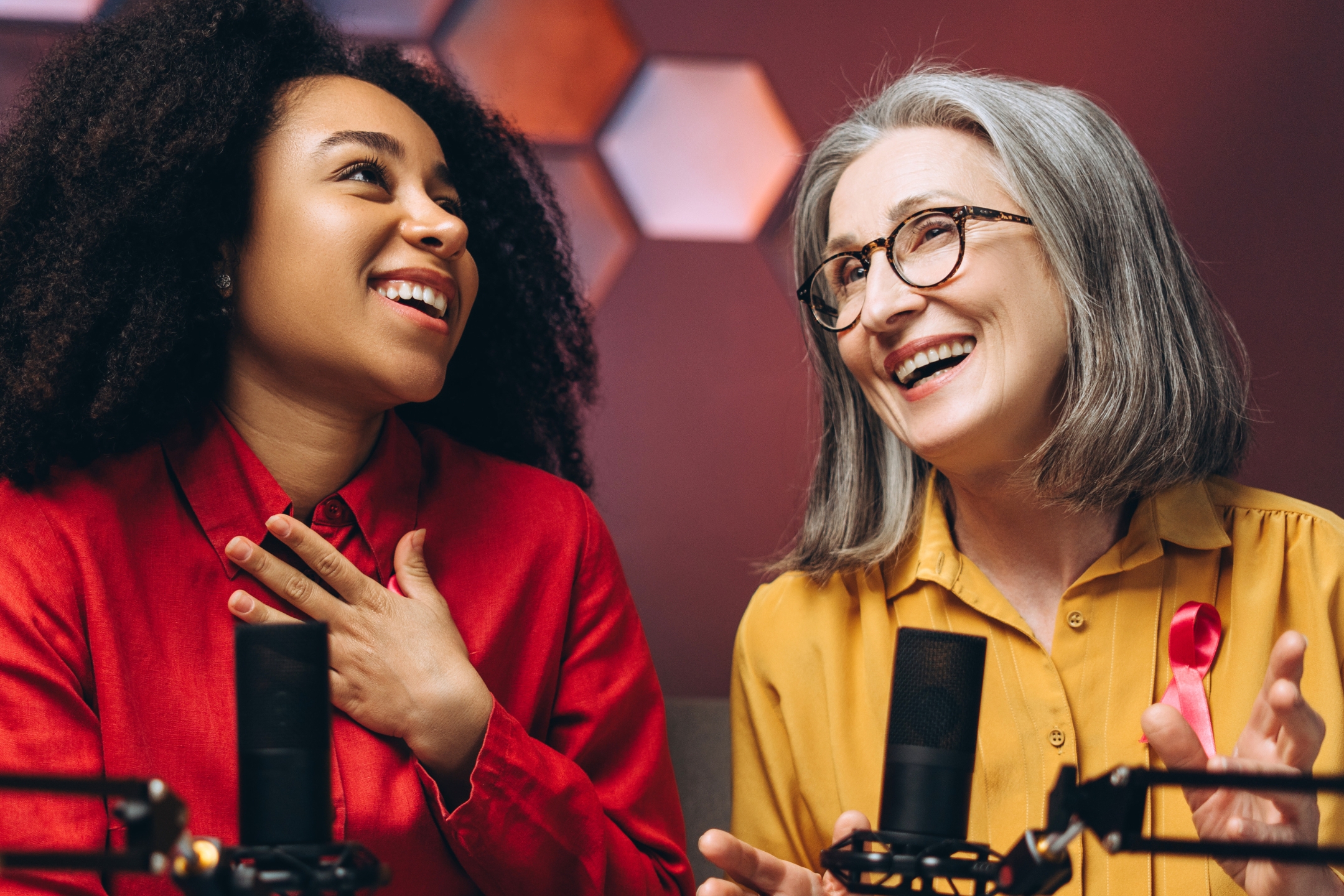 Two women laughing during a podcast recording in front of studio microphones